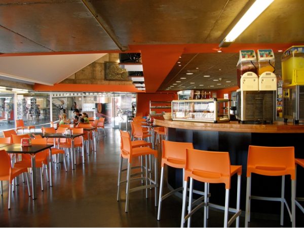 The image shows a modern cafe with orange bar stools and tables. There is a curved counter with slush machines and a display case. Large windows in the background let in natural light, and a few patrons are seated at tables. The interior has an industrial design.