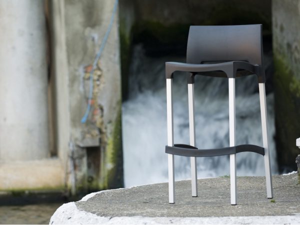 A modern bar stool with a black seat and white slender legs stands on a concrete surface beside a rushing waterway or waterfall. The background includes a weathered stone structure and flowing water.