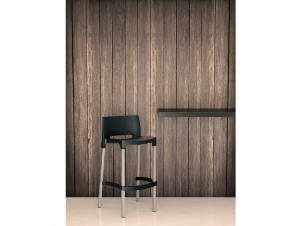 A modern black bar stool with metal legs is placed in front of a wooden panel wall. A small, minimalist floating shelf extends from the right side of the image. The floor is light-colored, creating a contrast with the dark tones of the stool and wall.