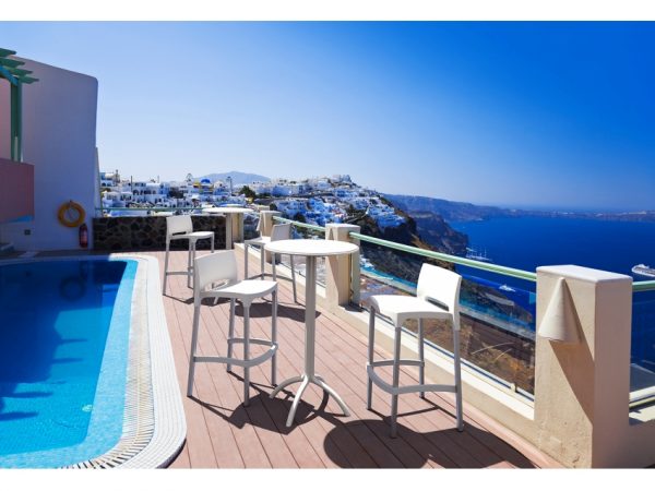 A rooftop deck with a small pool featuring a few tables and chairs. The area overlooks a picturesque coastal town with white buildings atop cliffs leading down to the deep blue sea below. The sky is clear and sunny, emphasizing the vibrant blue water.