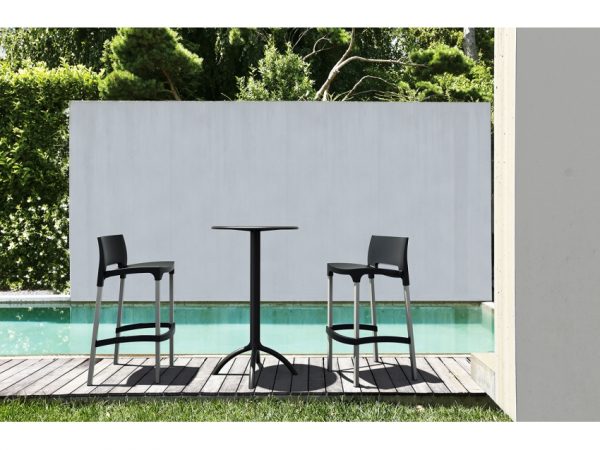 A modern outdoor seating area features a tall black table with two matching barstools on a wooden deck by a swimming pool. The setup is against a backdrop of lush green foliage and a white privacy wall. The scene evokes a minimalist, tranquil environment.