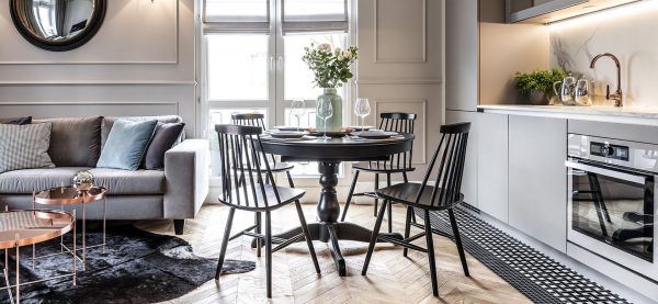 A modern, stylish living and dining area featuring a round black dining table accompanied by four 5910 Chairs. The space boasts gray paneled walls, a gray sofa adorned with cushions, and a kitchen outfitted with marble countertops. Decorative touches include a round mirror and a vase of flowers.