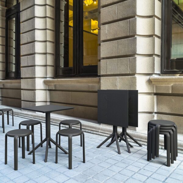 A street cafe scene with modern black tables and chairs on a light gray brick pavement. Some of the sleek Tom Stools are stacked nearby, along with several tables that are stacked upside down. The building behind features large windows and beige stone walls adorned with architectural detailing.